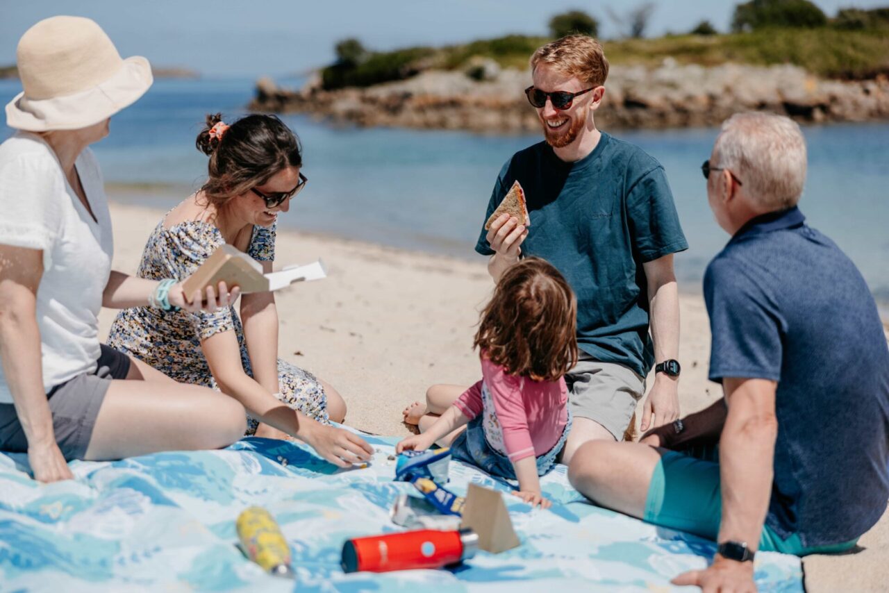 Family enjoying the beach, sat on a picnic blanket. 