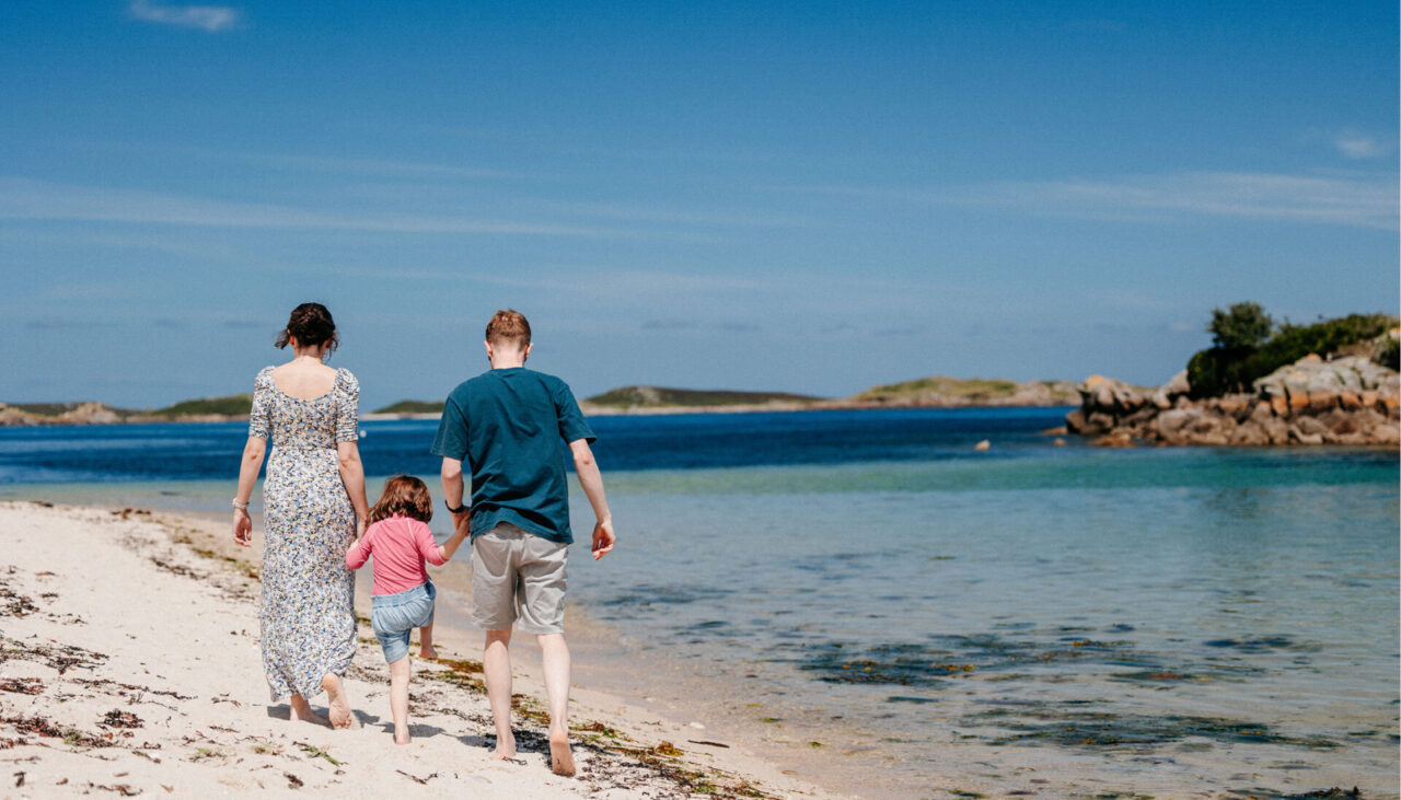 A family hand-in-hand walking across a beach on St Mary's Isles of Scilly, barefoot by the shoreline